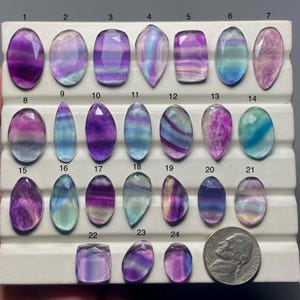 Peut inclure: Une collection de vingt-quatre pierres précieuses de fluorite polies, de formes variées, dont des ovales et des gouttes. Les pierres présentent une gamme de couleurs, principalement violet, vert et bleu, avec des motifs en bandes. Une pièce de monnaie américaine est incluse pour l'échelle.