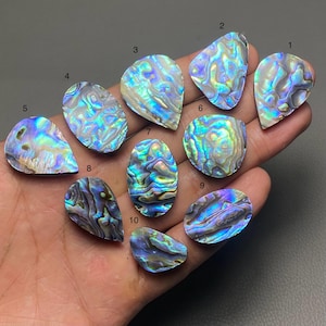 May include: A collection of ten iridescent abalone shell cabochons in various shapes, including oval, teardrop, and irregular forms. The shells display a vibrant array of colors, such as blue, green, and purple, with a pearlescent sheen. The cabochons are held in a hand.