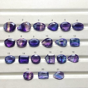 Könnte beinhalten: Eine Sammlung von einundzwanzig polierten Fluoritedelsteinen in verschiedenen Formen, darunter oval, tropfenförmig und rechteckig. Die Steine zeigen eine Mischung aus violetten, blauen und klaren Farbtönen. Jeder Stein ist nummeriert.