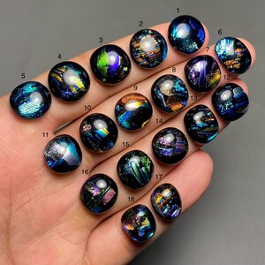 May include: Eighteen iridescent dichroic glass cabochons in various shades of blue, green, purple, and gold. The cabochons are arranged in rows on a hand.