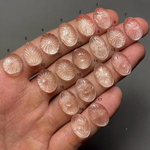Op de afbeelding: Een verzameling van achttien gesneden edelsteen cabochons in verschillende ovale en ronde vormen. Elke cabochon is een doorschijnende, licht perzikkleur en heeft een bloemen- of bladerontwerp. De edelstenen worden in een hand gehouden.