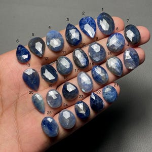 May include: A hand holds 25 blue sapphires, each with a unique cut and shape, including oval and teardrop forms. The gemstones vary in shade from light to dark blue. Each gem is numbered for reference. Measurements are in centimeters.