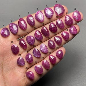 May include: A collection of twenty-nine faceted, oval-shaped, and teardrop-shaped ruby gemstones in varying shades of red and pink. Each stone is numbered, showcasing their unique cuts and clarity. The gemstones are displayed on a neutral background.