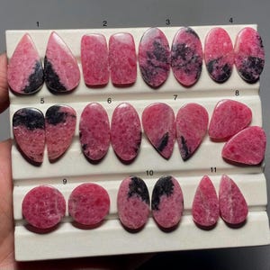 May include: A collection of eleven polished rhodonite gemstones in various shapes, including teardrop, oval, and rectangular. The stones display a vibrant pink hue with black veining, set against a white background.