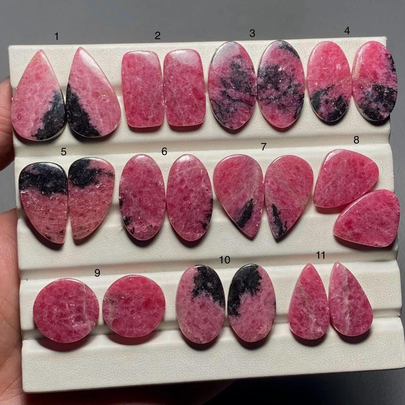 May include: A collection of eleven polished rhodonite gemstones in various shapes, including teardrop, oval, and rectangular. The stones display a vibrant pink hue with black veining, set against a white background.