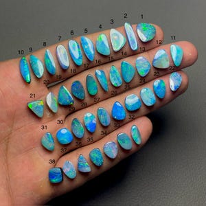 Puede incluir: Una colección de 38 ópalos de diversas formas y tamaños, mostrados en una mano. Los ópalos exhiben una gama de colores, incluyendo azules y verdes, con destellos iridiscentes. Cada ópalo está numerado.