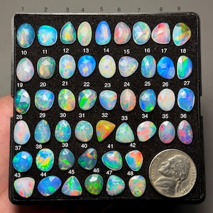 May include: A collection of 48 opals in various shapes and sizes, displayed in a black tray. The opals exhibit a range of colors, including blue, green, orange, and pink, with a nickel coin for size comparison. Each opal is numbered.