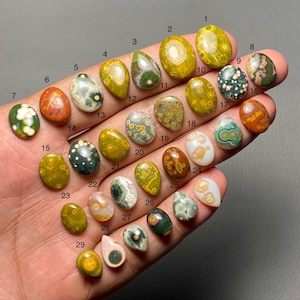 以下が含まれることがあります： さまざまな楕円形と涙滴型の29個の研磨された宝石のコレクション。石は、緑、黄、茶色、白など、さまざまな色を示し、ユニークなパターンとテクスチャがあります。宝石は手で持たれています。