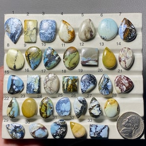 May include: A collection of thirty-three polished cabochon gemstones in various shapes and colors, including blue, yellow, white, and brown, arranged on a display tray with a US nickel coin for scale, each stone labeled with a number from 1 to 33.