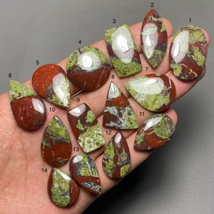 Könnte beinhalten: Sortiment von 14 roten, grünen und weißen Jaspis-Cabochons. Die Cabochons haben verschiedene Formen und Größen und sind auf einer Hand angeordnet.