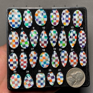 Op de afbeelding: Een verzameling van 27 regenboogkleurige glazen cabochons met een schaakbordpatroon in een zwarte displaycase. De cabochons zijn gerangschikt in rijen en kolommen, waarbij elke rij genummerd is van 1 tot en met 27. De cabochons zijn ovaal en traanvormig en hebben een verscheidenheid aan kleuren, waaronder blauw, groen, rood, oranje en geel.