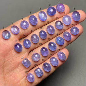 May include: Twenty-seven oval-shaped gemstones, numbered 1 through 27, in shades of blue and purple, arranged in rows on a hand.