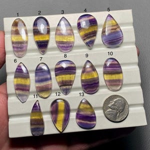 May include: Thirteen purple, yellow, and white striped fluorite cabochons in a teardrop shape, arranged in rows on a white display board. A US penny is included for scale.