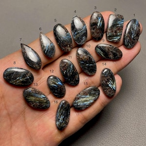 May include: Sixteen polished, oval and teardrop-shaped dark gemstones with iridescent blue and metallic streaks, each labelled with a small black number from 1 to 16. The stones exhibit a fibrous, chatoyant texture with deep brown and black base colours.