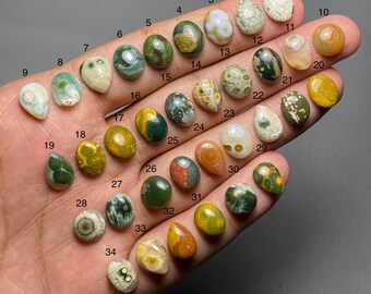May include: A selection of 34 polished gemstone cabochons in diverse shapes and colours, including green, brown, yellow, and white. Each stone is numbered, displaying the variety of natural patterns and textures. The gemstones are held in a hand.
