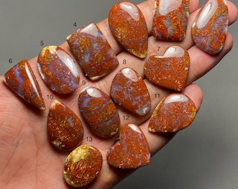 May include: A selection of polished gemstones in diverse shapes, such as oval, rectangular, and heart-shaped. The stones exhibit a blend of red, orange, and purple tones with distinctive patterns. The gemstones are held in a hand.