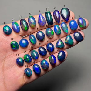 Peut inclure: Une collection de trente opales polies de formes et tailles variées, présentées sur une main. Les opales présentent une gamme de couleurs, notamment des bleus, des verts et des noirs, avec des reflets irisés. Chaque opale est numérotée.