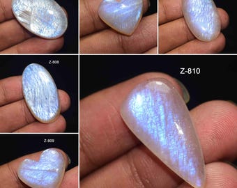May include: A collection of polished moonstone gemstones in various shapes, including oval, heart, and teardrop. The stones display a shimmering, iridescent effect with blue and white hues. Each stone is labeled with a "Z" code.