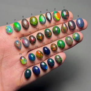 May include: A hand displays thirty oval-shaped opals, each with a unique color and pattern. The opals range in color from green, blue, red, and orange, with some displaying iridescent qualities. Each stone is numbered from 1 to 30.