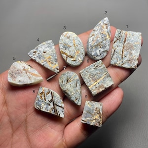 Op de afbeelding: Negen witte en grijze edelsteen cabochons met gouden vlekken. De cabochons zijn in verschillende vormen en maten.