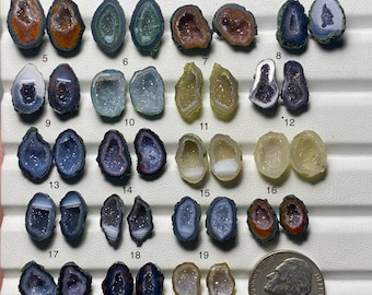 May include: A collection of nineteen pairs of small, polished agate geode slices arranged in rows on a white display tray, numbered one through nineteen. Each pair features unique crystalline centers in shades of orange, blue, purple, green, and yellow, with a US nickel coin included for scale.