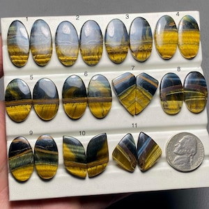 May include: Twelve pairs of oval and teardrop shaped blue and gold tiger's eye cabochons, arranged in rows on a white display card. The cabochons are approximately 1 inch in length.