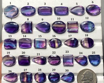 May include: A collection of thirty faceted fluorite gemstones in various shapes including oval, pear, square, and rectangular cuts. The stones display purple, blue, and clear color banding. A US nickel coin is placed next to the stones for scale.