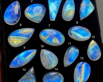 May include: A collection of fifteen polished moonstone gemstones, each with a unique shape and iridescent blue and yellow hues. The stones are displayed on a black surface, showcasing their natural beauty and variations.