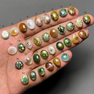 以下が含まれることがあります： 緑、茶色、白、黄色など、さまざまな色と模様の34個の研磨された宝石のコレクション。石は楕円形で、手の上に表示され、独特の質感とデザインが示されています。各石には番号が付けられています。