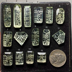 Puede incluir: Una colección de tallas de piedras preciosas verde claro, cada una con intrincados diseños florales. Las formas varían, incluyendo rectángulos, óvalos y un diamante. Se incluye una moneda de cuarto de dólar estadounidense para referencia de tamaño. Las piedras están sobre una superficie negra.