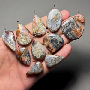 May include: A collection of ten polished agate gemstones in various shapes and sizes. The stones display a range of colors, including orange, gray, white, and brown, with intricate banding patterns. Each stone is unique, showcasing natural variations.