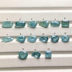 Puede incluir: Una colección de quince gemas de aguamarina en varias formas, incluyendo cortes rectangulares, de pera y ovalados. Las gemas son de color azul claro y están dispuestas sobre una superficie blanca. Cada gema está numerada.