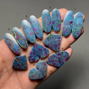 May include: A collection of polished cabochon gemstones featuring a vibrant mix of blue, green, and magenta-pink inclusions. The stones are shaped into ovals, triangles, and teardrops, each marked with a small black number from one to nine.