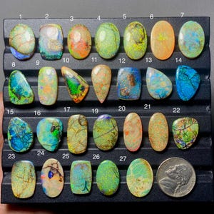 May include: A collection of 27 polished opal gemstones in various shapes and colors, including oval, teardrop, and rectangular. The opals display a range of colors, such as green, blue, orange, and yellow, with unique patterns and internal structures. A US nickel is included for size comparison.