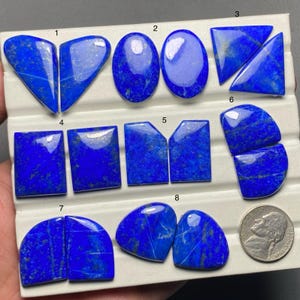 Könnte beinhalten: Acht Paare blauer Lapislazuli-Cabochons in verschiedenen Formen und Größen, darunter Herz, Oval, Rechteck und unregelmäßige Formen. Die Cabochons sind in Reihen auf einer weißen Oberfläche angeordnet und von 1 bis 8 nummeriert.