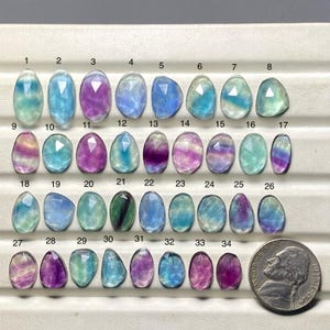 May include: A collection of 34 faceted fluorite gemstones in various shapes and colors, including purple, blue, green, and yellow. A US nickel coin is included for size comparison. The gemstones are arranged on a white surface.