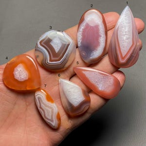 Op de afbeelding: Zeven agaat cabochons in verschillende vormen en maten, met een mix van oranje, roze en witte banden. De cabochons zijn genummerd van 1 tot en met 7.