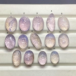 Könnte beinhalten: Eine Sammlung von vierzehn geschnitzten Edelsteinen in verschiedenen ovalen und blattförmigen Formen. Die Edelsteine sind hauptsächlich lavendelfarben und weiß, mit floralen Designs, die in ihre Oberflächen geschnitzt sind.