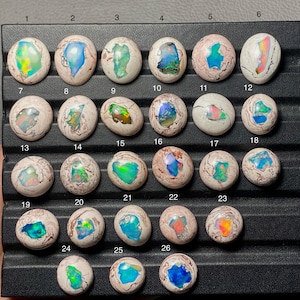 May include: A collection of twenty-six oval-shaped opal gemstones, each with unique internal patterns of color. The opals display a variety of colors, including blue, green, pink, and yellow, set against a creamy white or tan base.
