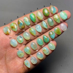 Op de afbeelding: Een verzameling van dertig ovale en druppelvormige opalen in verschillende tinten groen, blauw en geel. De edelstenen worden op een hand getoond, waarbij hun unieke kleuren en patronen worden benadrukt. Elke steen is genummerd.