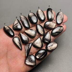 Puede incluir: Diecinueve cabujones de piedra preciosa negra y plateada, cada uno con un patrón y una forma únicos, están dispuestos en una mano. Los cabujones están numerados del 1 al 19.