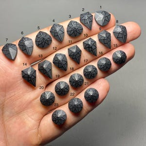 May include: A collection of twenty-three gray gemstone cabochons with geometric black designs. The cabochons vary in shape, including teardrop, diamond, and round, and are numbered 1 through 23.