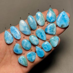 May include: A collection of 17 blue and white gemstone cabochons, numbered 1 through 17, arranged on a hand. The cabochons are various shapes and sizes, with a pearlescent, iridescent finish.