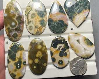 Könnte beinhalten: Neun polierte Ozeanjaspis-Cabochons in verschiedenen Formen und Größen, darunter oval, tropfenförmig und herzförmig. Die Cabochons weisen eine Vielzahl von Farben auf, darunter Grün, Braun, Weiß und Gelb. Die Cabochons sind auf einer weißen Oberfläche mit den Zahlen 1 bis 8 angeordnet.