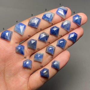 May include: A selection of faceted blue gemstones in diverse geometric forms, such as squares, diamonds, and pentagons. The gemstones are presented on a hand, highlighting their colour and cut. The gemstones are numbered from 1 to 18.