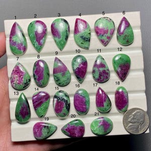 Peut inclure: Vingt et un cabochons de pierres précieuses vertes et roses, numérotés de 1 à 21, disposés en rangées sur un panneau d'affichage blanc. Une pièce de 10 centimes d'euro est incluse pour l'échelle.