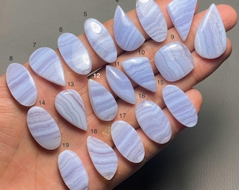 Puede incluir: Una colección de diecinueve piedras preciosas de ágata de encaje azul pulidas en varias formas, incluyendo lágrimas y óvalos. Cada piedra muestra un patrón de bandas de tonos azul claro y blanco, con un acabado liso y brillante. Las piedras preciosas están numeradas del 1 al 19.