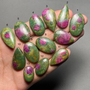 May include: A collection of thirteen polished ruby gemstones, each with a unique shape and pattern. The rubies display a vibrant pink-red hue, set against a green and gold matrix. The gemstones vary in size and shape, suitable for jewelry making.