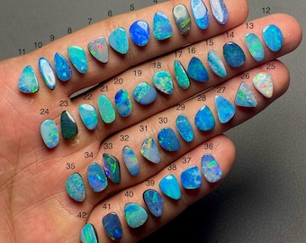 Puede incluir: Una colección de 42 piedras preciosas de ópalo de varias formas y tamaños, mostradas en una mano. Los ópalos exhiben una gama de colores, incluyendo azules, verdes y destellos iridiscentes. Cada piedra está numerada.