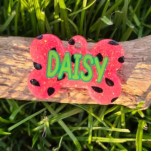 May include: Pink and black bone-shaped dog tag with the name "DAISY" written in green glitter.